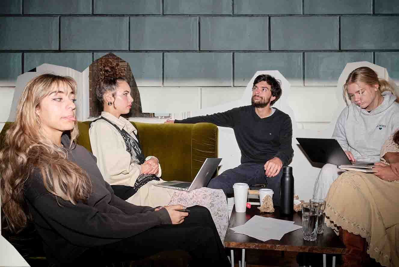 Four individuals are seated on and around a green couch in a casual meeting setting, engaged in a discussion with laptops and coffee cups present.