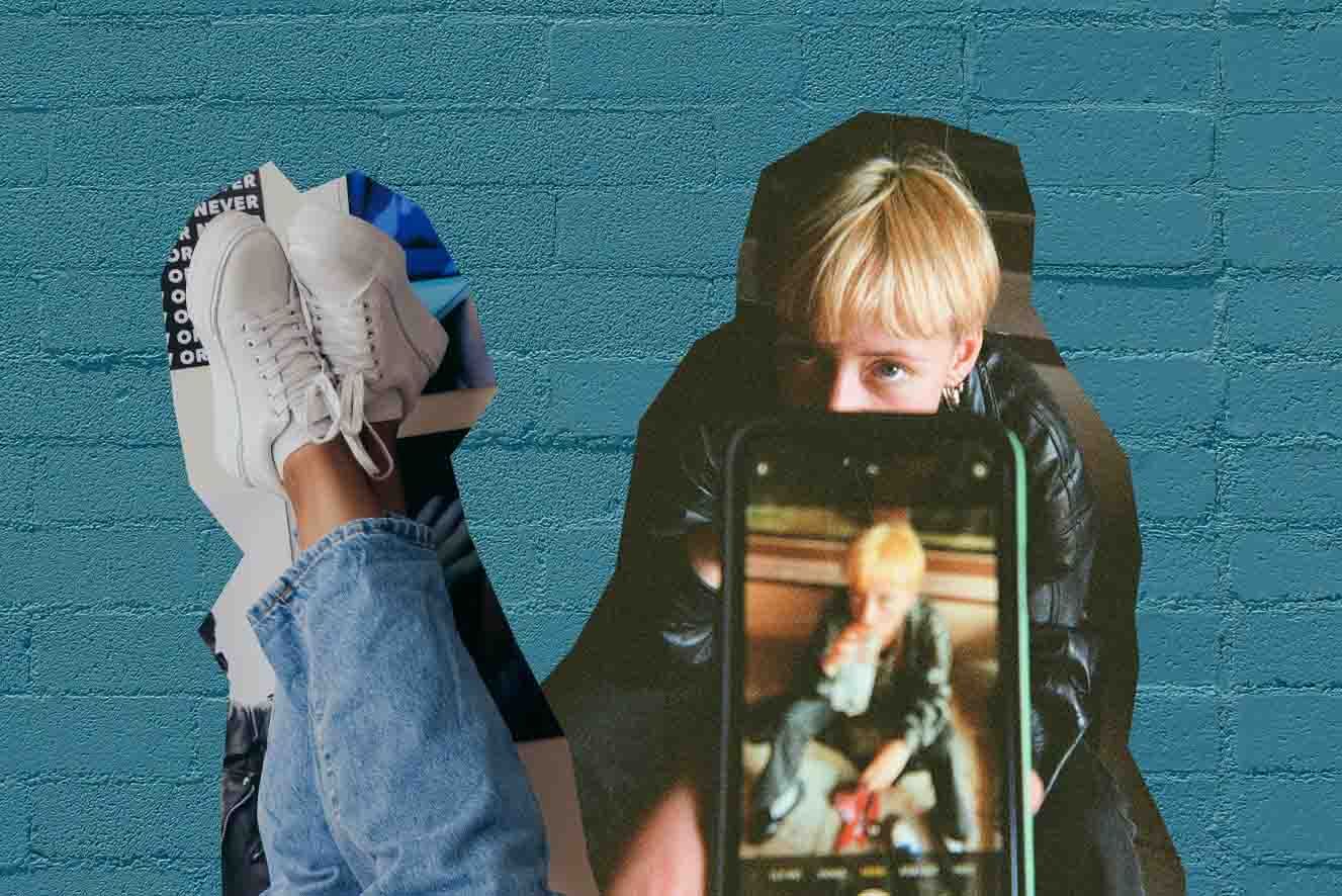 A person is taking a selfie with a smartphone. The phone screen visible in the image displays the selfie being taken.  To the left is sneaker-clad feet against a wall painted blue