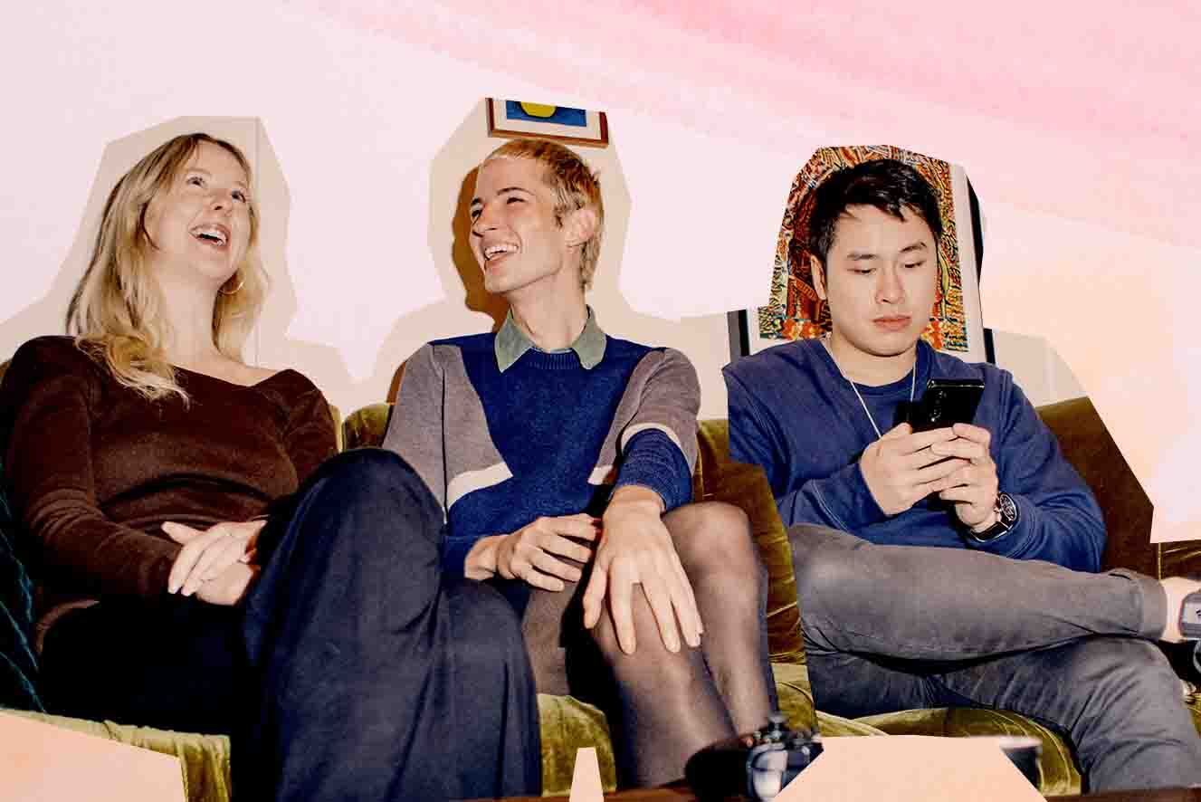 Three individuals sitting together; one is laughing, another smiling and looking away, and the third focused on a mobile phone. They are sitting in a casual indoor setting.
