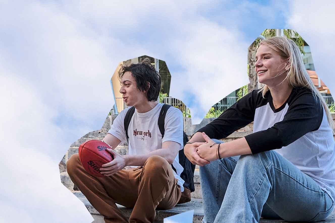 A young man and women sit side by side. In front of a blue sky bacground The male wears a backpack and holds a red football. The female has long blonde hair and wears a black and white t-shirt with blue jeans